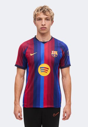 Joven vestido con la camiseta de fútbol a rayas rojas y azules del FC Barcelona con los logos de Nike y Spotify, de pie frente a un fondo claro y liso.