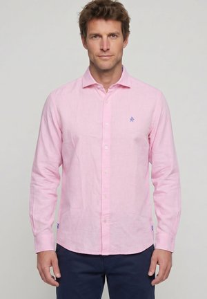 Hombre con cabello corto y castaño que lleva una camisa de botones color rosa claro y pantalones oscuros, de pie frente a un fondo claro y liso.