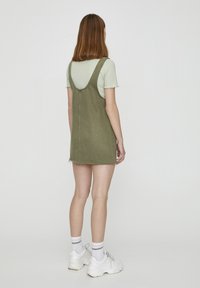 Robe en denim vert olive avec un corsage ajusté et une jupe évasée, dotée d'un dos en col rond et d'un ourlet effiloché. Associée à une chemise rayée et des baskets.