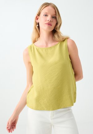 Jonge vrouw met blond haar draagt een mouwloos lichtgroen topje en witte broek, poseert tegen een effen lichte achtergrond.