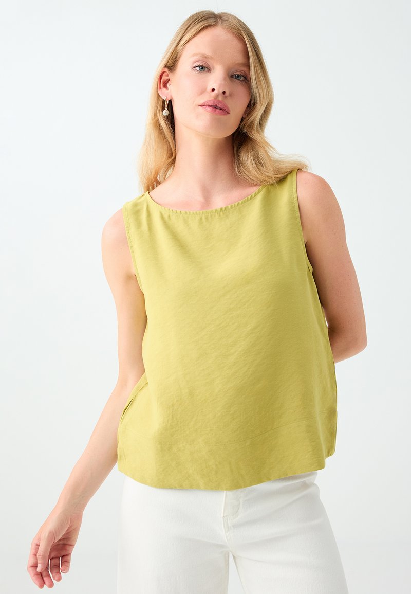 Jeune femme aux cheveux blonds porte un haut sans manches vert clair et un pantalon blanc, posant devant un fond clair uni.