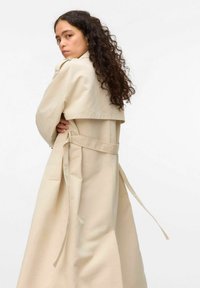 Beige trenchcoat med bälte i midjan, breda slag och en flödande design. Har en dubbelknäppt front och långa ärmar.