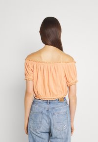 Blusa arancione a spalle scoperte con bordi arricciati, tessuto testurizzato e vita arricciata, abbinata a jeans in denim chiaro rovinosi.