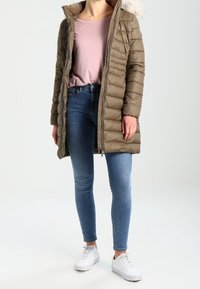 Abrigo acolchado ligero en verde oliva con capucha de piel sintética, combinado con una blusa rosa claro, jeans ajustados azules y zapatillas blancas.