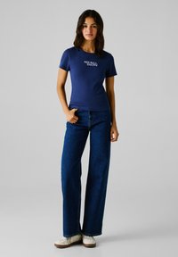 Camiseta de manga corta de canalé en color marino con el logo "RED BULL RACING", combinada con pantalones vaqueros de pierna ancha en azul y zapatillas blancas.