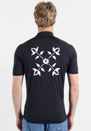 Homme aux cheveux blonds courts vu de dos, portant un t-shirt noir à manches courtes avec un motif floral blanc et feuillu dans le dos.