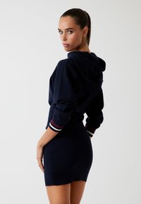 Rochie cu glugă de culoare bleumarin, realizată dintr-un material moale. Dispune de mâneci lungi cu manșete în dungi și o siluetă ajustată. Design minimalist.