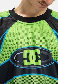 Zaļš un melns īsām piedurknēm svārku krekls ar acu tekstūru, lielu DC logo un zilām akcentiem ar punktu rakstiem.