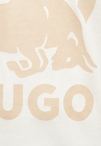 Λογότυπο ταύρου με υφή μπεζ πάνω από τα μεγάλα γράμματα «UGO» σε λευκό φόντο από ύφασμα.