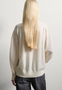 Pull oversized beige clair avec une texture douce, épaules tombantes et poignets côtelés ; porté avec un jean gris foncé. Vue arrière.