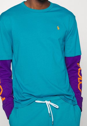 Türkises T-Shirt mit kurzen Ärmeln und lilanen langen Ärmeln. Verfügt über ein orangefarbenes Logo auf lila, aus Baumwollmaterial und im Rundhalsausschnitt-Design.