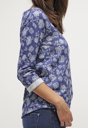 Mujer con una camisa de manga larga azul con patrones florales blancos y puños de encaje, combinada con vaqueros azules, mostrada de perfil.