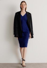 Mørkeblå kjole med en tettsittende silhuett, kombinert med en svart oversized blazer. Har stroppeholdte svarte hæler og minimale tilbehør.