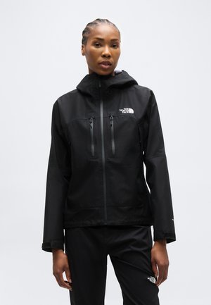 Femme portant une veste et un pantalon noirs The North Face, debout devant un fond clair uni, faisant face à l'avant avec une expression neutre.