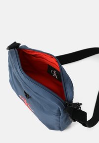 Bolso de hombro de tela azul con cierre de cremallera. Forro rojo en el interior, con un parche de logo. Correa negra ajustable y tirador de cremallera de metal.
