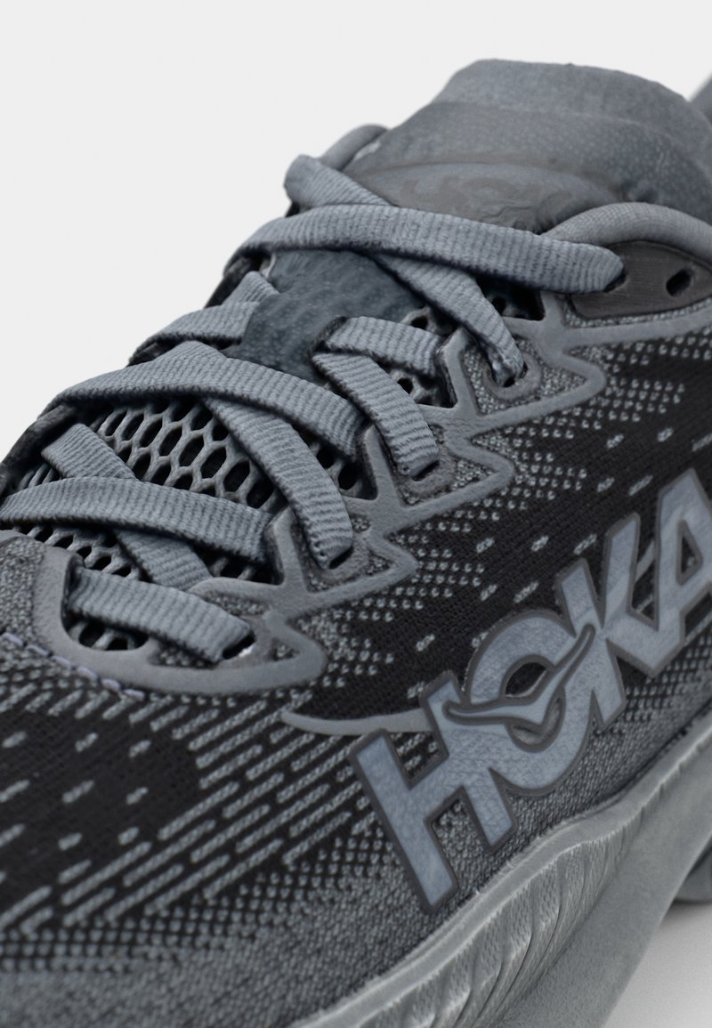 Grauer Sportschuh mit einem Obermaterial aus Mesh, strukturiertem Design, flachen Schnürsenkeln und einem markanten Logo an der Seite. Gepolsterte Zwischensohle mit schlankem Profil.