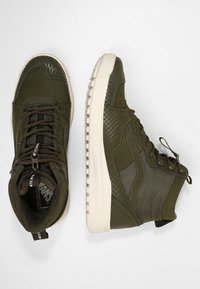 Zapatillas altas de color verde oliva con paneles texturizados, costuras tonales y una suela de goma blanca. Presentan cordones y una etiqueta con el logotipo en la lengüeta.