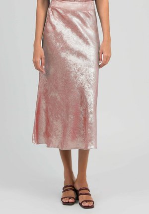 Jupe midi rose clair scintillante portée par une personne debout, associée à des sandales à talons marron à brides sur fond uni.