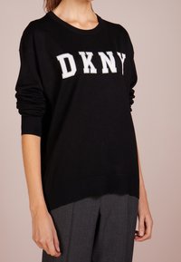 Osoba v černém svetru DKNY s bílým nápisem a šedých kalhotách, stojící před jednobarevným světlým pozadím.