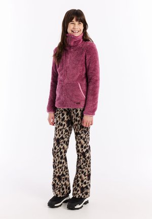 Glückliches Mädchen in einem lila Fleecejacke mit Reißverschluss, Leoprint-Hose und schwarzen Stiefeln, stehend vor einem weißen Hintergrund.