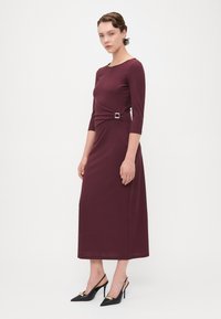 Mauve midi šaty s trojštvrťovými rukávmi, ktoré majú stočený predný detail a akcent zlatého štvorcového spona. Skombinované s čiernymi lodičkami s špicatou špičkou.