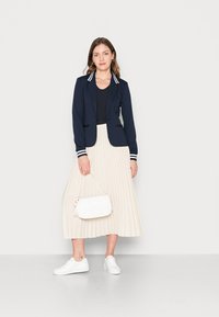 Blazer blu navy con colletto a righe, top nero, gonna plissettata beige, sneakers bianche e borsa bianca trapuntata.