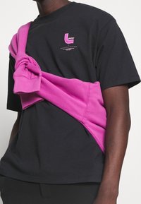 T-shirt en coton noir avec logo imprimé rose, accompagné d'un sweat-shirt rose noué autour des épaules, accentuant le contraste des couleurs et les superpositions.