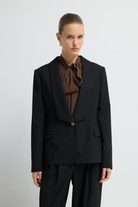 Žena s hladkými vlasy svázanými dozadu, nosící stříbrné kruhové náušnice, hnědou blůzu s mašlí a černý blazer s proužky a odpovídající kalhoty.