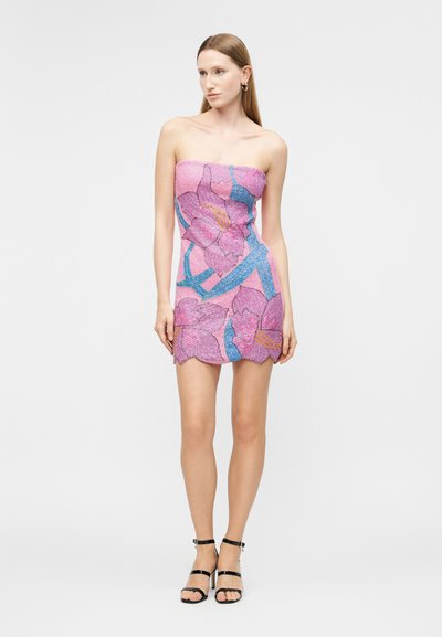 Ženska oblečena v kratko roza obleko brez naramnic z velikimi vijoličastimi cvetovi in modrimi poudarki, obuta v črne sandale z naramnicami.