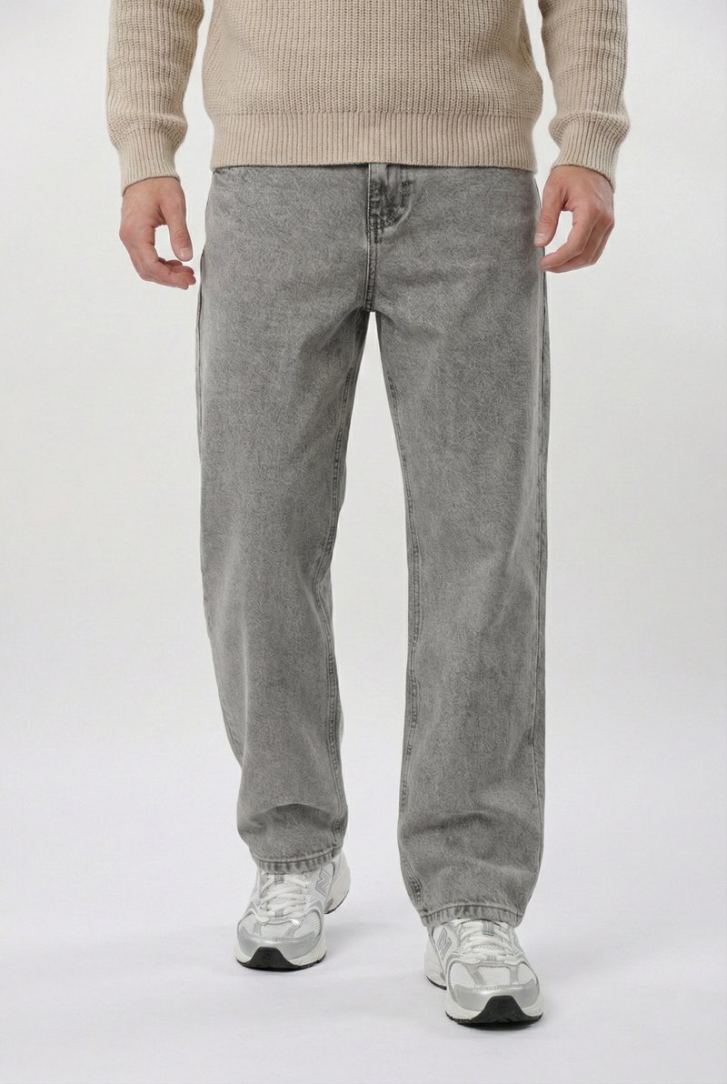 Mann trägt hellgraue, locker sitzende Jeans, beige gestrickten Pullover und weiße Turnschuhe, steht vor weißem Hintergrund.