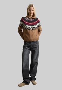 Pull en laine marron avec un empiècement multicolore, associé à un jean foncé et des chaussures beige clair. Présente une coupe décontractée et un ourlet côtelé.