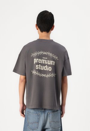 T-shirt oversize en coton gris à manches courtes, avec le texte "premium studio" et des graphiques décoratifs de feuilles dans le dos.