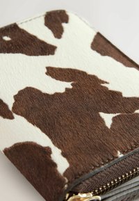 Barna és fehér tehénbőr tárca, puha, texturált felülettel, cipzárral és barna varrással az élein.