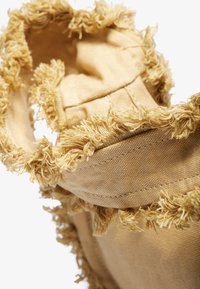 Beigefarget stoffprodukt med teksturerte, nupperte kanter. Myk, lodden trim gir detaljer rundt åpningen og fremhever den unike designen og materialet.