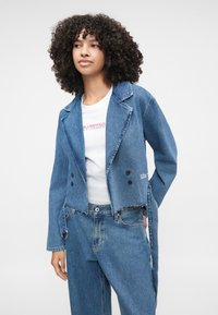 Izgriezta zila džinsa jaka ar izliektu apkakli, divu pogu aizdari, nolietotu apakšdaļu un sānu saiti, valkāta virs baltas T-krekla.