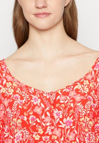 Blusa vermelha floral com decote redondo, apresentando padrões de flores rosas e amarelas, mangas curtas e fechos de botão à frente.