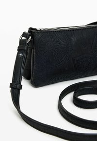 Sac bandoulière en cuir noir avec des motifs circulaires embossés, une fermeture éclair en métal argenté et une sangle réglable. Texture lisse avec une forme structurée.