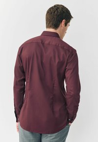 Bordeaux langærmet skjorte med skræddersyet design, glat tekstur, knapærmer og en klassisk krave, båret over grå bukser.