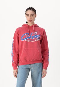 Rød hettegenser med frontlomme, blå grafisk tekst "Chaos Custom Supply", og blå stjerneakenter på ermene; myk stoffkvalitet og avslappet passform.
