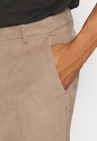 Beige manchesterbyxor med texturerad yta, utrustade med sidofickor och en rak skärning, visas med en hand i en ficka.