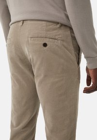 Beżowe spodnie z korduroju z pionowymi żeberkami, tylne kieszenie z guzicznymi akcentami oraz zwężanym krojem, widok z tyłu.