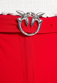 Boucle de ceinture en argent en forme de deux oiseaux face à face sur une ceinture rouge vif portée avec un haut blanc texturé.