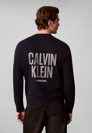 Homme de dos portant un sweat-shirt noir avec CALVIN KLEIN et Established 1968 imprimés en grandes lettres dans le dos.
