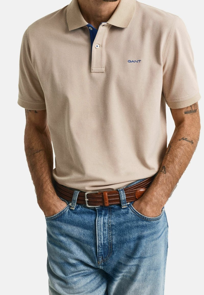 Polo beige avec un col bleu, portant un logo sur la poitrine, accompagné d'un jean en denim bleu et d'une ceinture en cuir tressé marron.