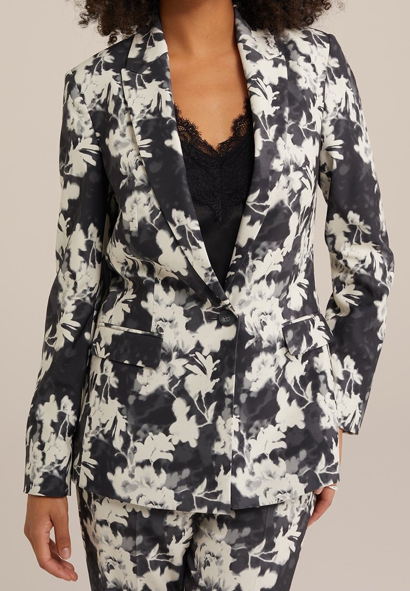 Femme portant un blazer à motif floral noir et blanc assorti à un pantalon, avec un haut noir bordé de dentelle, debout devant un fond neutre.