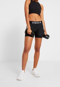 Svart sportig crop top och shorts med "UNDER" midjeband, håller hantlar, kombinerat med vita sportskor, mot en vit bakgrund.
