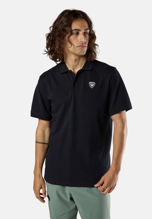 Mann mit schulterlangem, lockigem Haar, der ein schwarzes Polohemd mit kleinem Logo auf der Brust und hellgrüne Hosen trägt und zur Seite schaut.