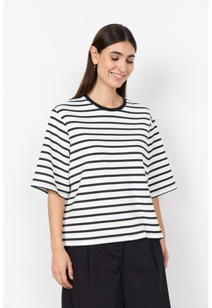 Vrouw die glimlacht, draagt een wit T-shirt met korte mouwen met zwarte horizontale strepen en zwarte wijde broek, staand tegen een witte achtergrond.