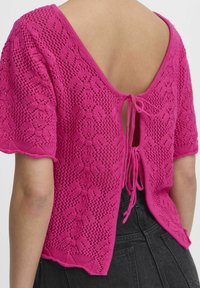 Adīta blūze, dzīvīgā rozā krāsā, ar teksturētu rakstu, īsām piedurknēm un atvērtu aizmuguri, kas aizverama ar auklām.