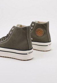 Zapatillas altas verde oliva con suelas gruesas blancas, una raya negra, ojales metálicos y un parche redondo marrón con el logo en el lateral.